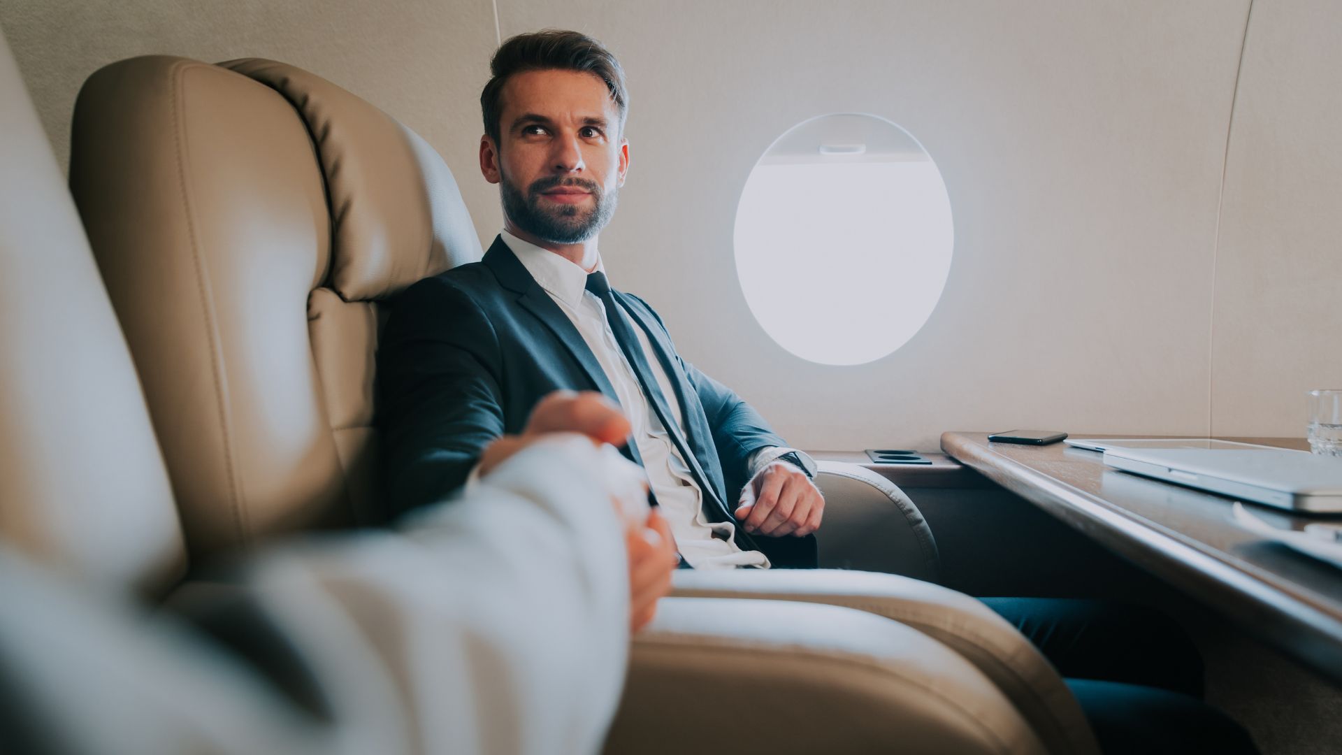 A man in a suit sits in a private jet, shaking hands with another person as they discuss consent preferences. A laptop and glass of water are on the table beside him.