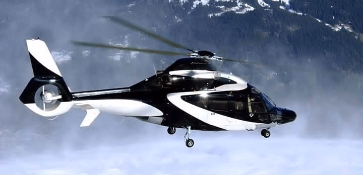 A black and white helicopter flies above snow-covered ground with mountains and trees visible in the background.