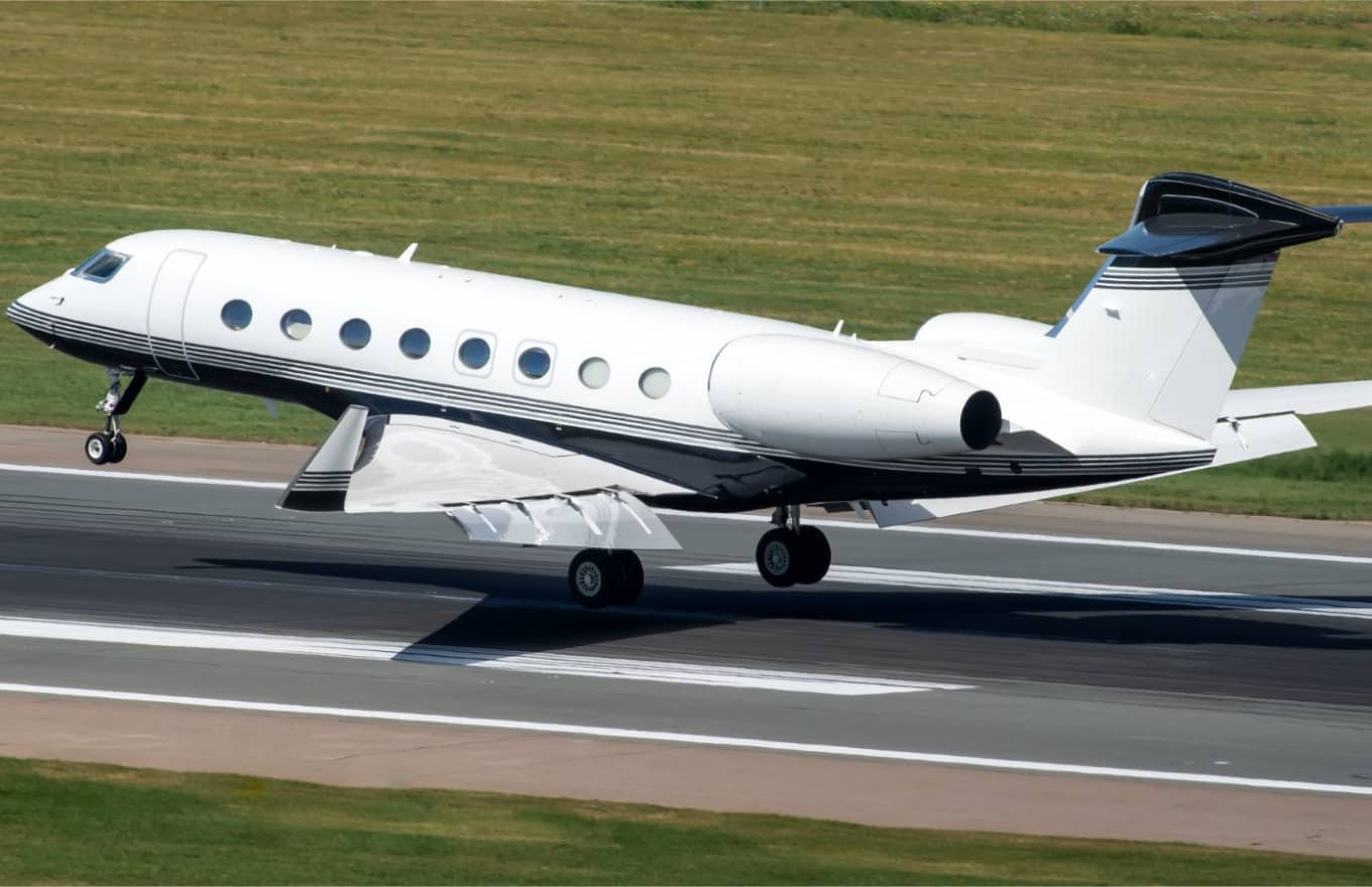 A white private jet with six windows is landing on a runway with a grassy field in the background.