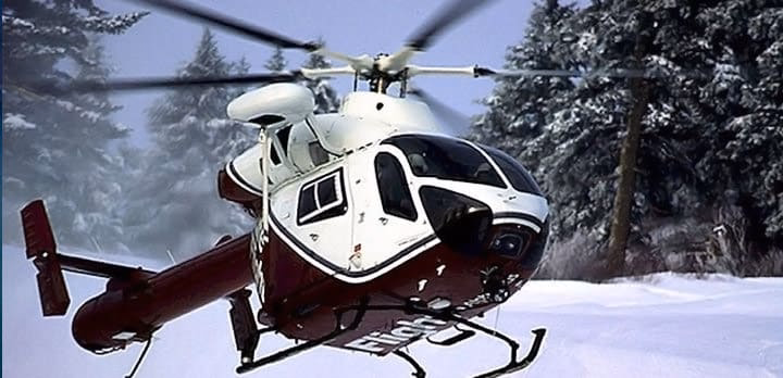 A red and white rescue helicopter flies low over snow-covered ground with tall, snow-laden trees in the background.