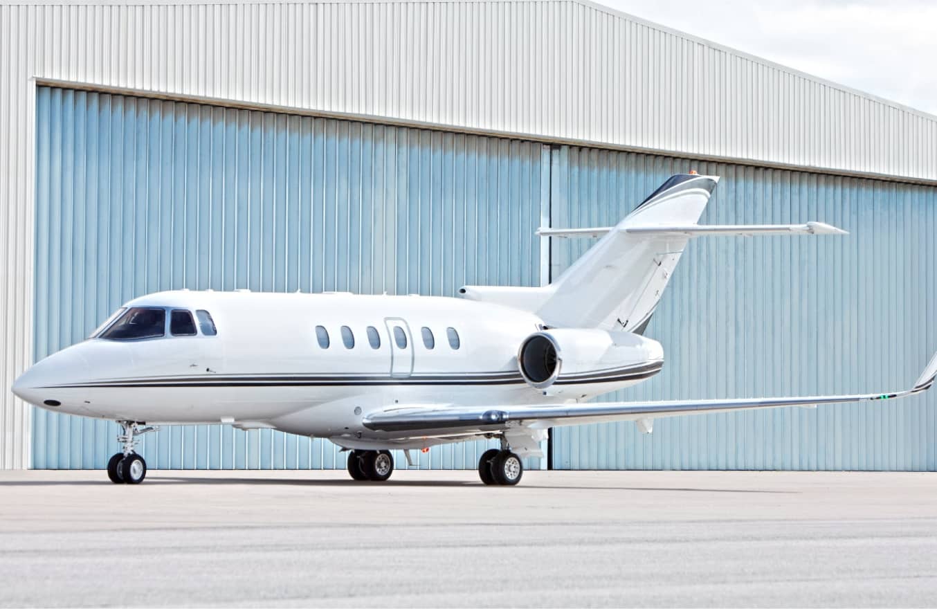 A white private jet is parked on the tarmac in front of a large corrugated metal hangar.