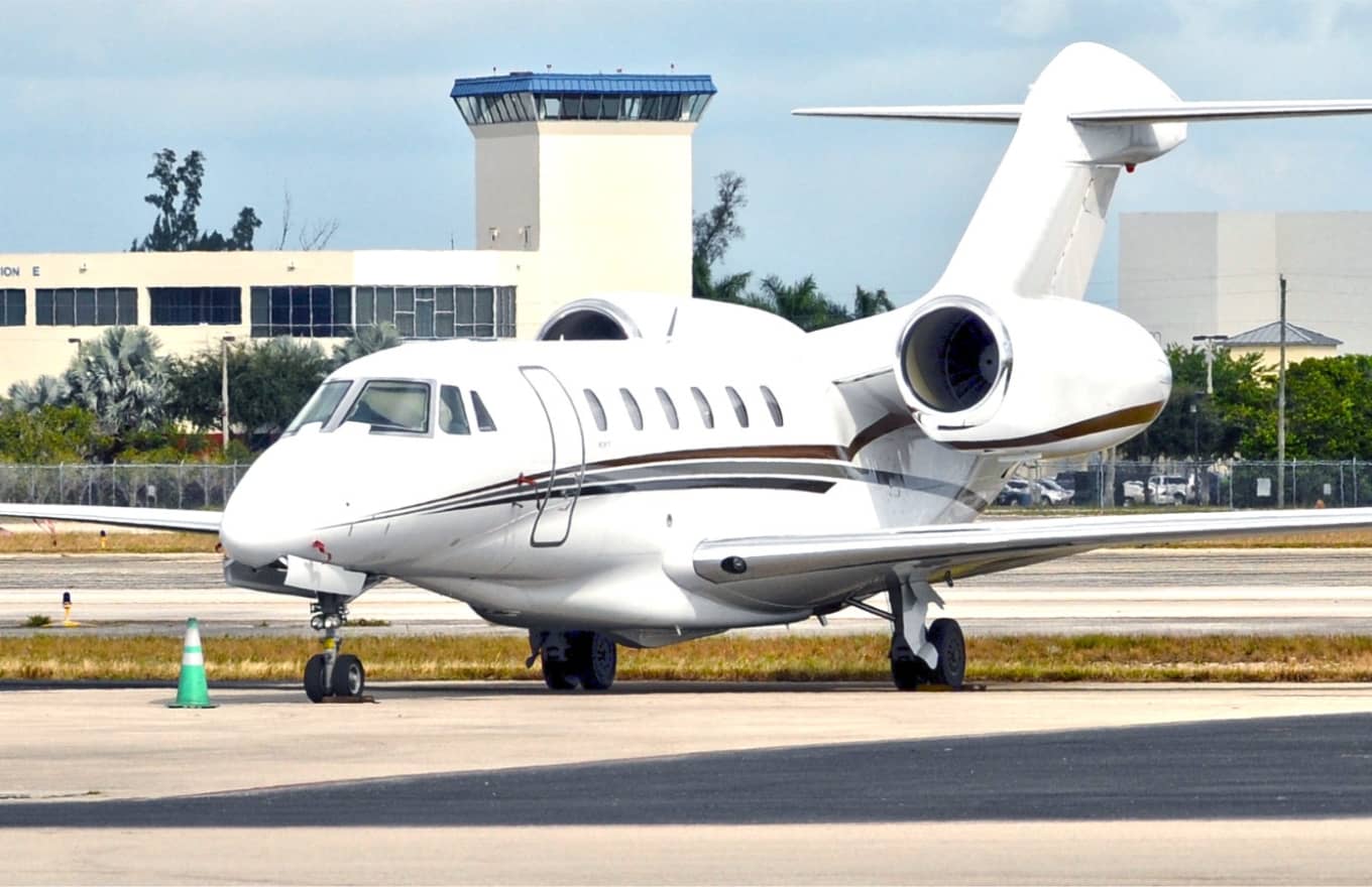 A white private jet is parked on an airport runway with a control tower and several buildings in the background.