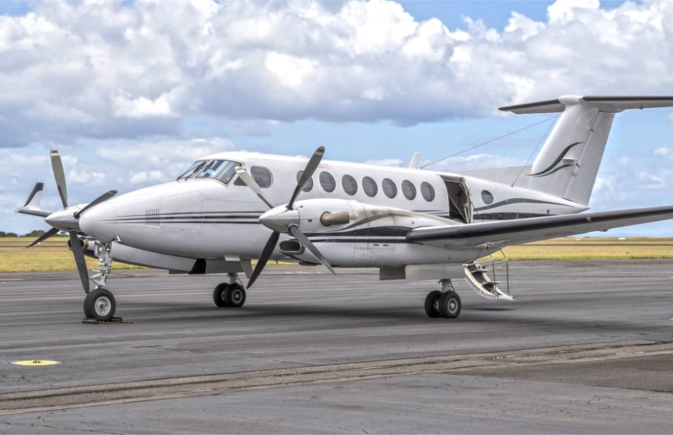 A white twin-propeller aircraft is parked on an airport tarmac with its door open under a partly cloudy sky.
