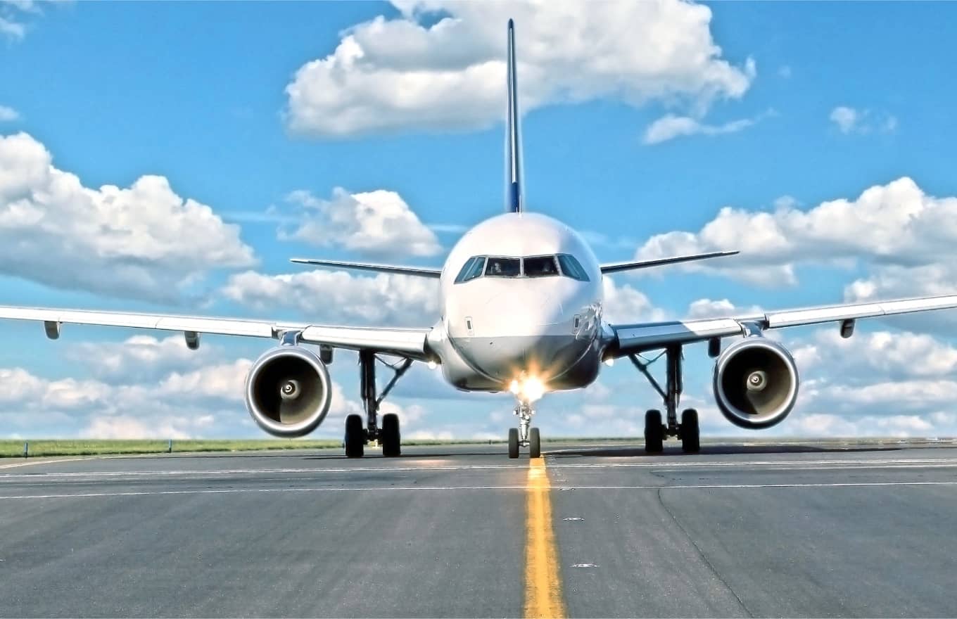 A commercial airplane is positioned on a runway under a blue sky with scattered clouds, facing directly toward the camera.