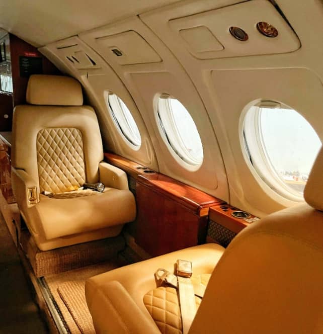 Interior of a private jet with two tan leather seats, seatbelts fastened, and three oval windows on the side.