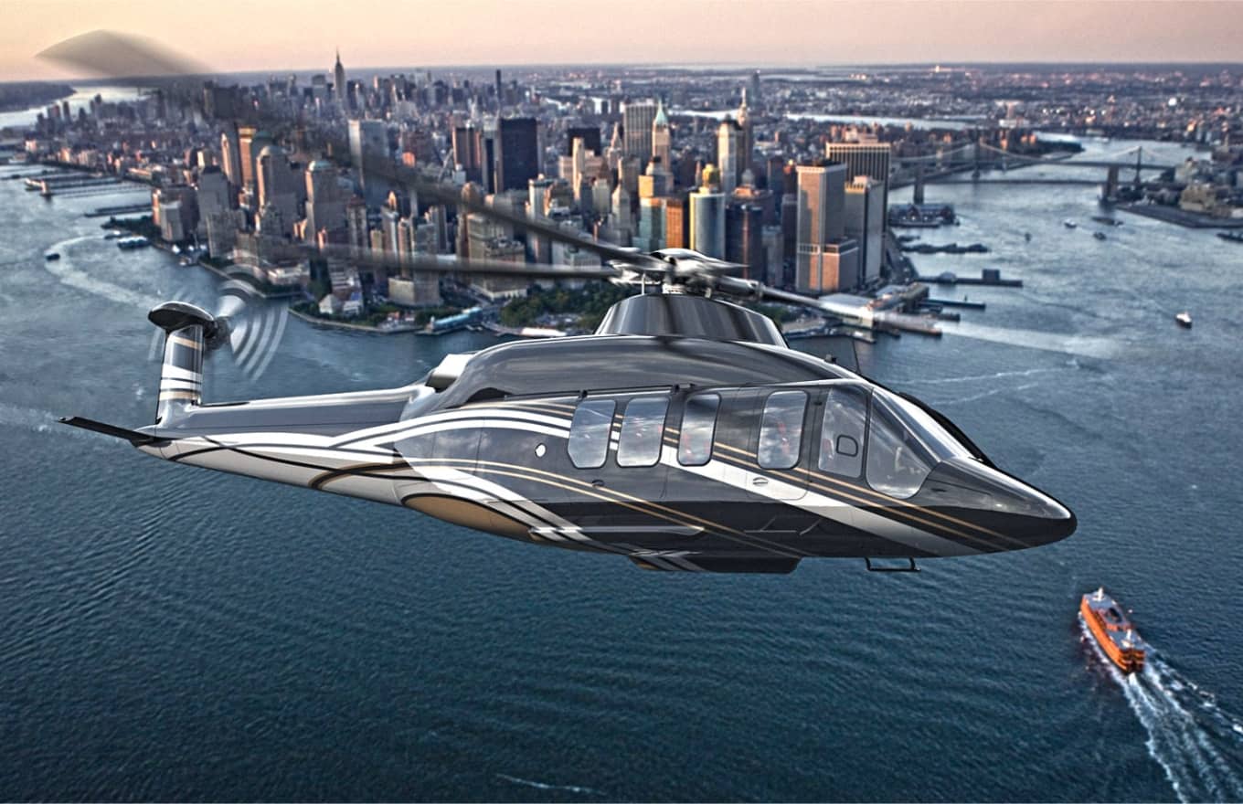 A sleek black and silver helicopter flies over a river with the New York City skyline and a ferry visible in the background at sunset.