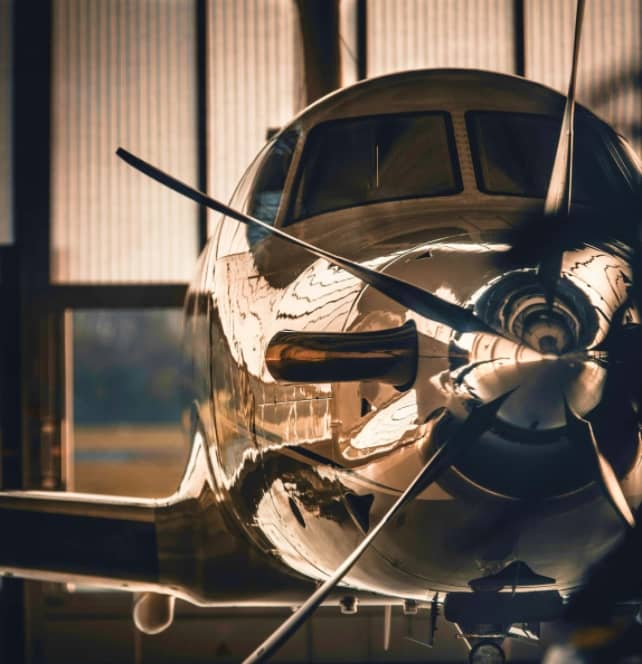 Close-up view of a propeller airplane nose and cockpit, reflecting light, with the propeller blades in focus inside a hangar.